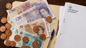 Top-down photograph of stacked British banknotes and copper coins alongside an HMRC-branded document inside a folder on a dark walnut table.